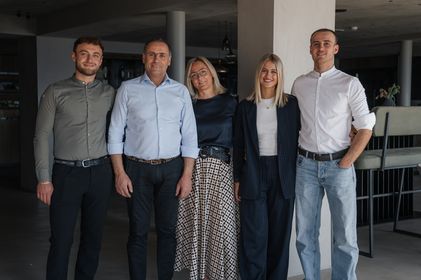 Eine Gruppe von fünf Personen, drei Männer und zwei Frauen, steht in einem modern gestalteten Innenraum nebeneinander und lächelt in die Kamera.