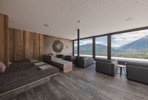 Ein moderner Ruheraum mit Holzwand, einer großen Liegefläche und mehreren grauen Sesseln vor einer Fensterfront mit Panoramablick auf die Alpen.