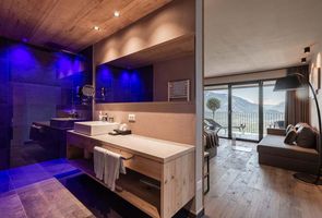 Ein modernes Hotelzimmer mit offenem Badezimmer, das durch eine Holzwand vom Wohnbereich getrennt ist; links ein Waschbereich mit blauem Licht und Spiegel, rechts ein gemütliches Zimmer mit Blick auf die Berge.