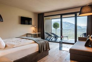 Modernes Hotelzimmer mit Doppelbett, Sofa, Fernseher und bodentiefen Fenstern, die auf einen Balkon mit Bergblick führen.