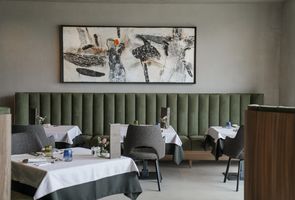 Ein Restaurant-Innenraum mit einer langen, grünen gepolsterten Sitzbank vor einer Wand mit einem großen abstrakten Kunstwerk. Die Tische sind mit weißen Tischdecken und Gedecken vorbereitet.