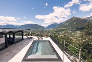Eine moderne Terrasse mit Swimmingpool, Sonnenliegen und Blick auf ein weitläufiges Alpental mit Bergen unter blauem Himmel.