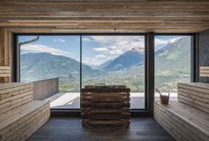 Ein Blick aus dem Inneren einer hölzernen Sauna auf ein sonniges Bergpanorama mit einem Tal im Hintergrund durch ein großes Panoramafenster.